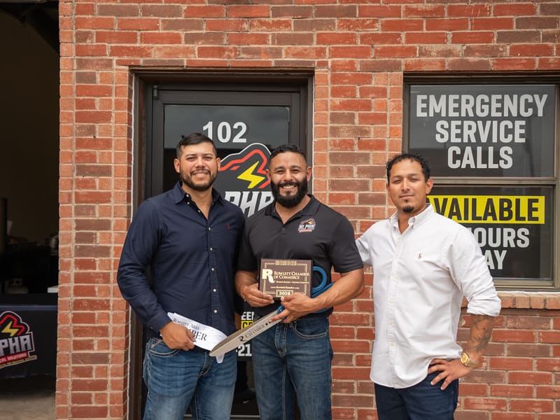 Three men outside Alpha Electrical's Rowlett office beside an Emergency Service Calls sign, one holding a chamber plaque