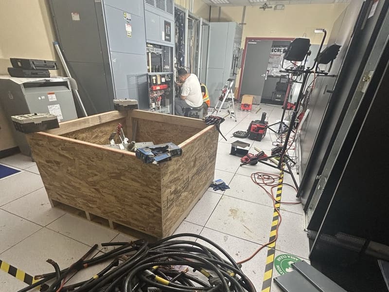Technicians work at open switchgear in an electrical room, with a wooden crate of parts and a pile of cut cable in front