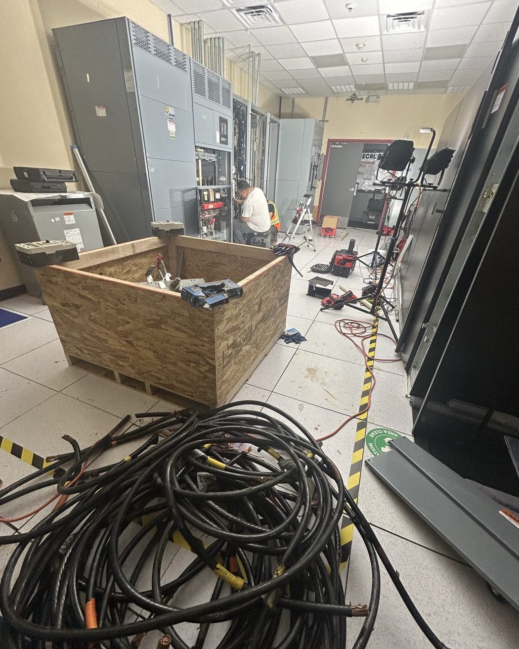 Technicians work at open switchgear in an electrical room, with a wooden crate of parts and a pile of cut cable in front