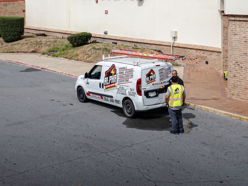 White Alpha Electrical van with a roof ladder parked at a retail store entrance, two workers beside it, one in a safety vest