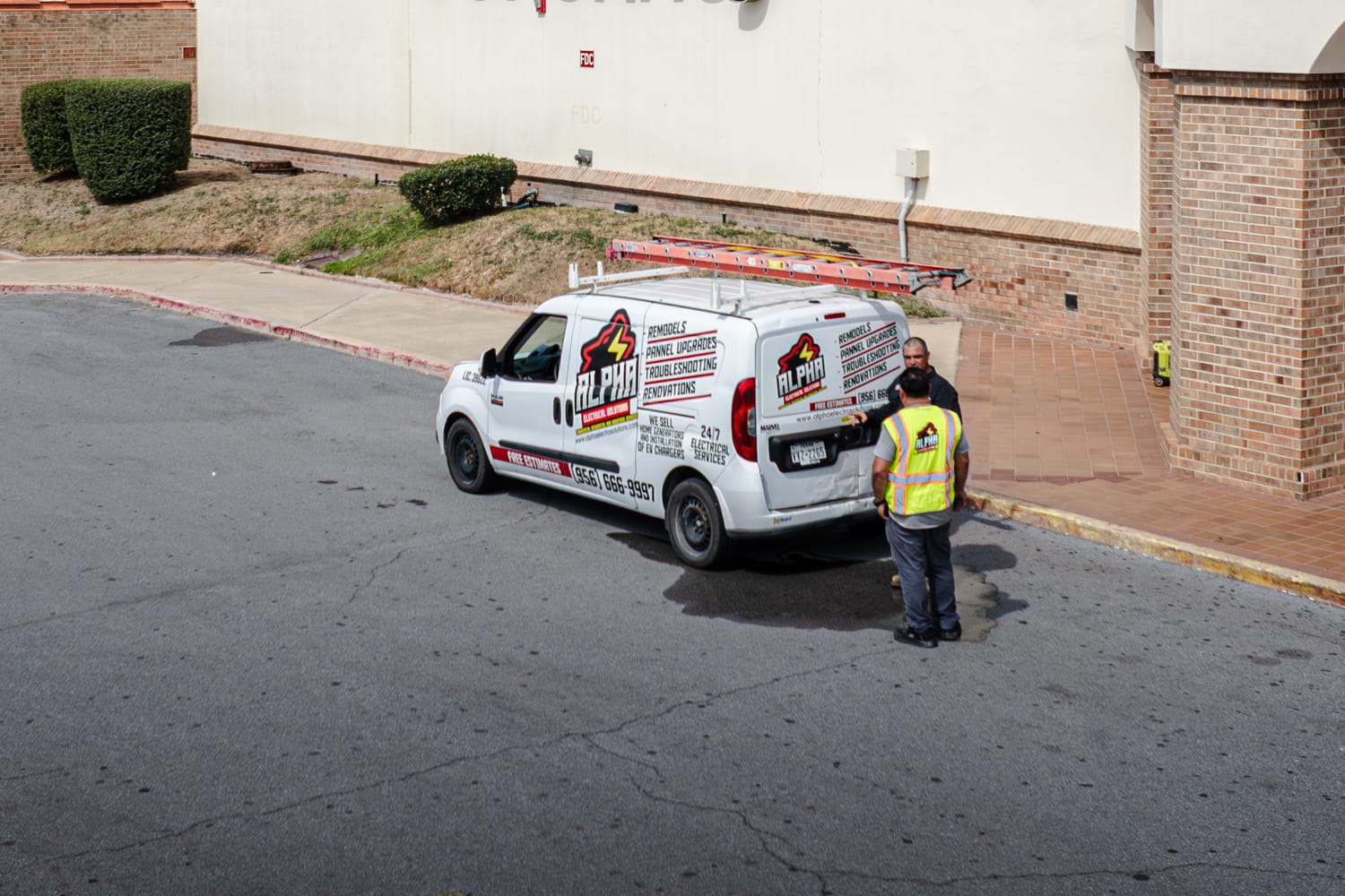 White Alpha Electrical van with a roof ladder parked at a retail store entrance, two workers beside it, one in a safety vest