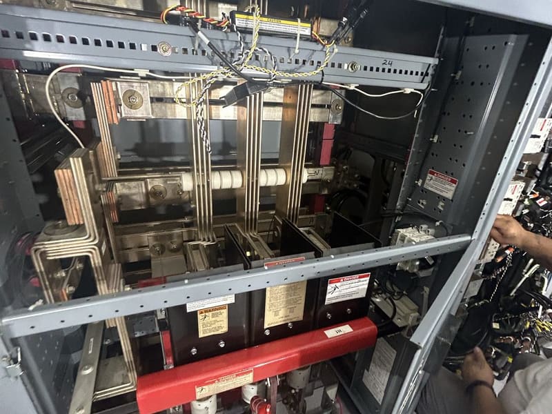Open gray switchgear cabinet showing bus bars and a large breaker with warning labels, a technician working at right