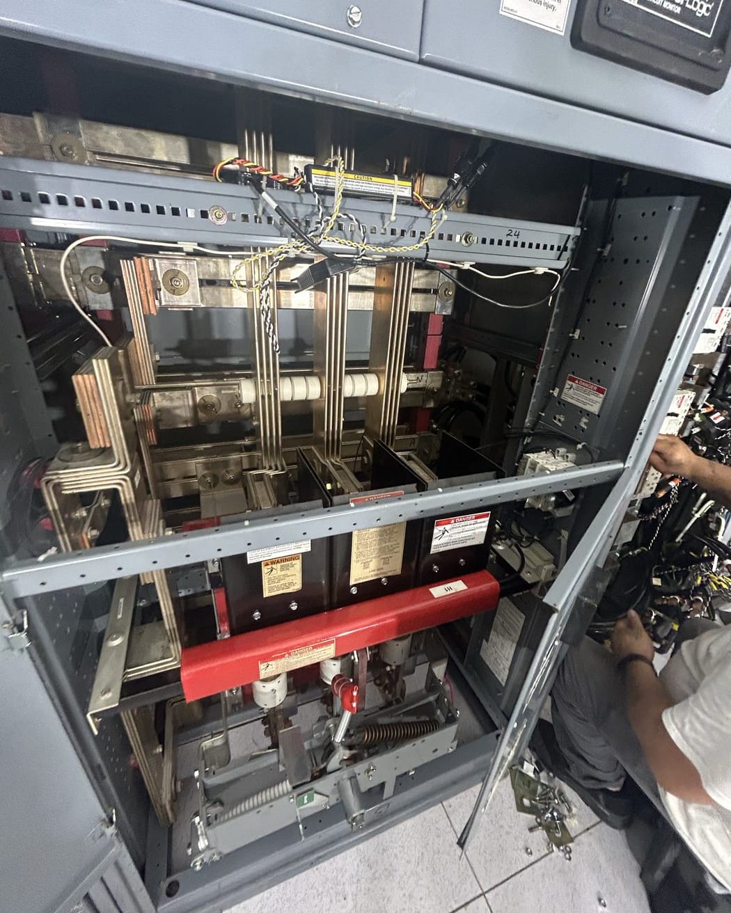 Open gray switchgear cabinet showing bus bars and a large breaker with warning labels, a technician working at right