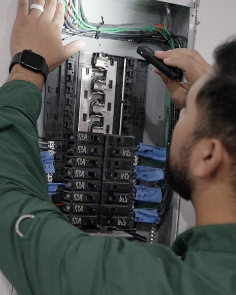 Technician in a green shirt points a handheld tool at an open home breaker panel with blue tape labels on the circuits