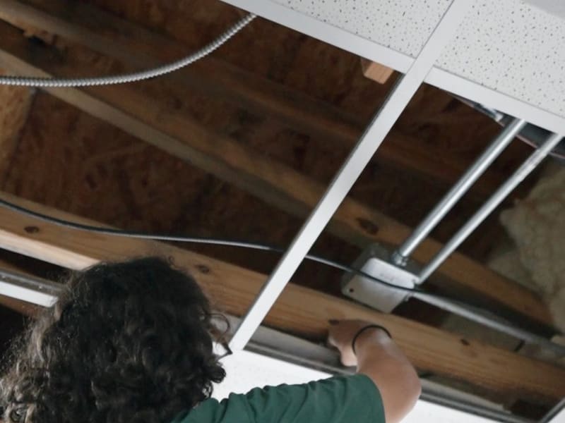Electrician on an orange ladder reaches into an open drop ceiling where conduit and a junction box run along wood framing
