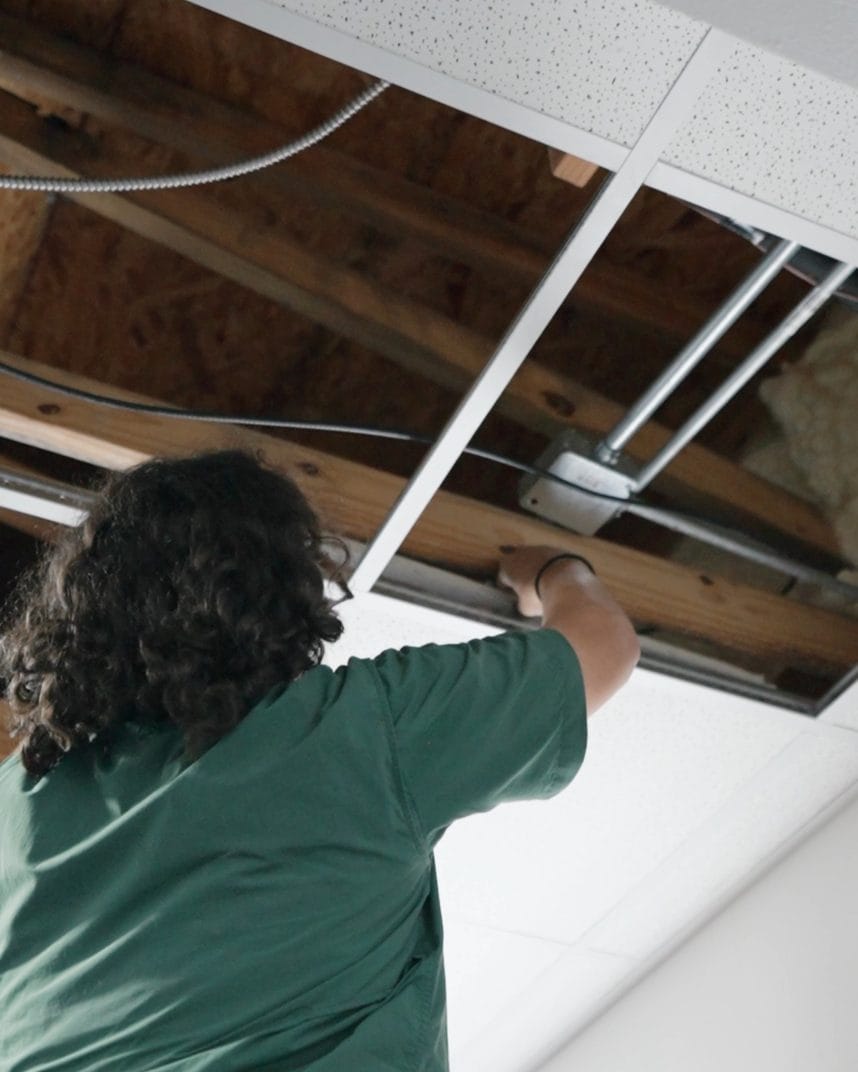 Electrician on an orange ladder reaches into an open drop ceiling where conduit and a junction box run along wood framing